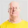 A bald man with glasses wearing a yellow shirt looks directly at the camera against a plain light background.