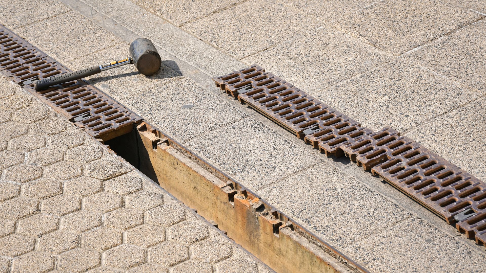 A rubber mallet rests on pavement next to a partially open metal drainage grate, revealing a gap in the ground.