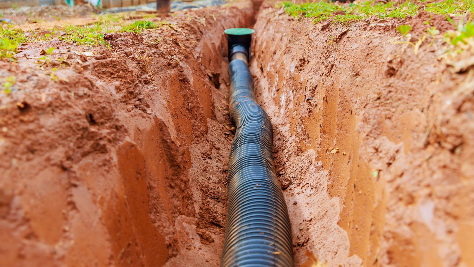 A large black drainage pipe is installed in a narrow trench dug into reddish-brown soil, partially surrounded by grass.