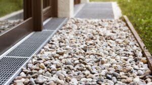 Close-up view of a gravel bed with a metal drainage grate next to the exterior wall of a building. Grass is visible at the edge of the gravel.