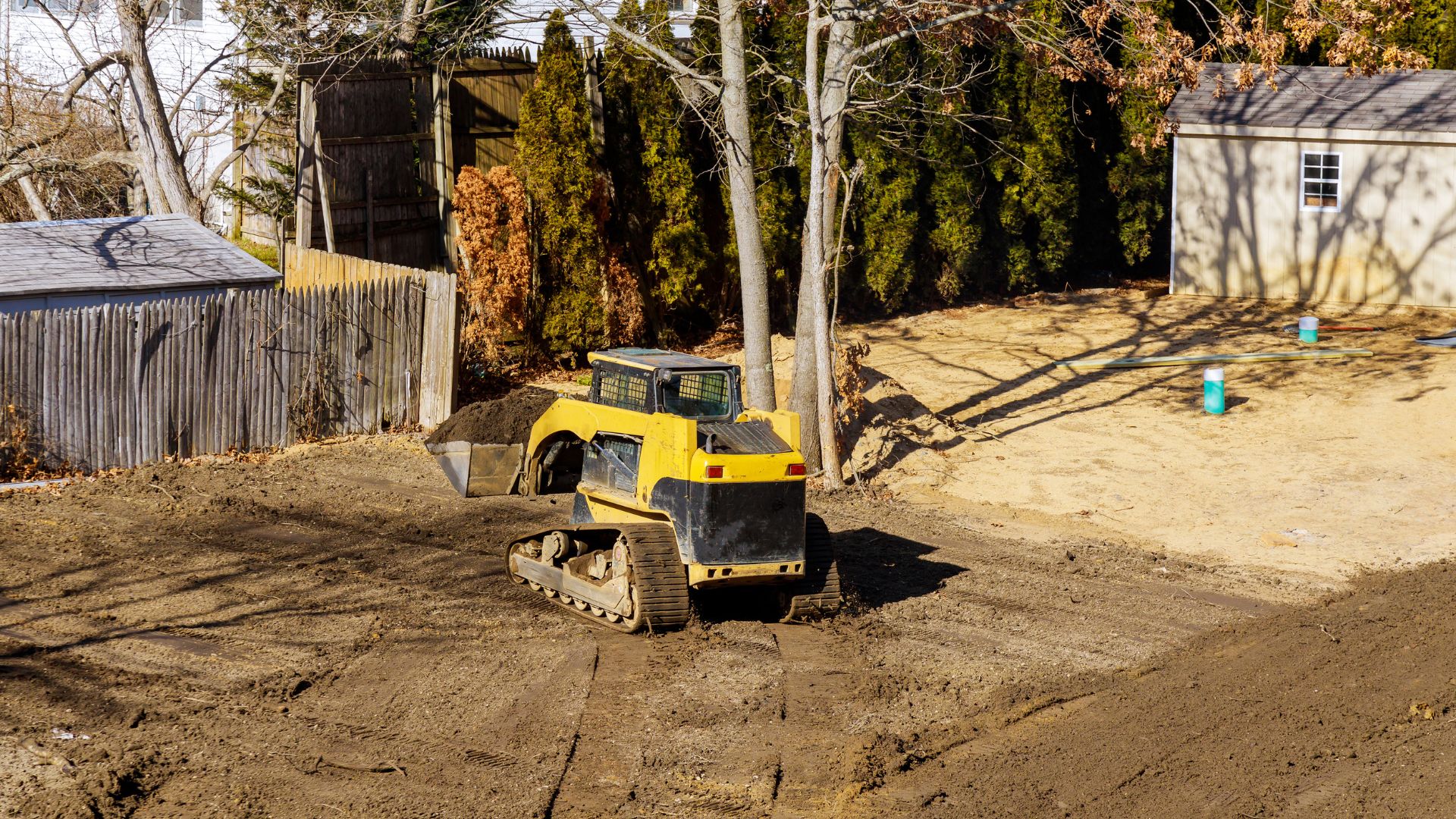 A yellow bulldozer is moving dirt in a backyard with bare trees, a wooden fence, and a small shed nearby.